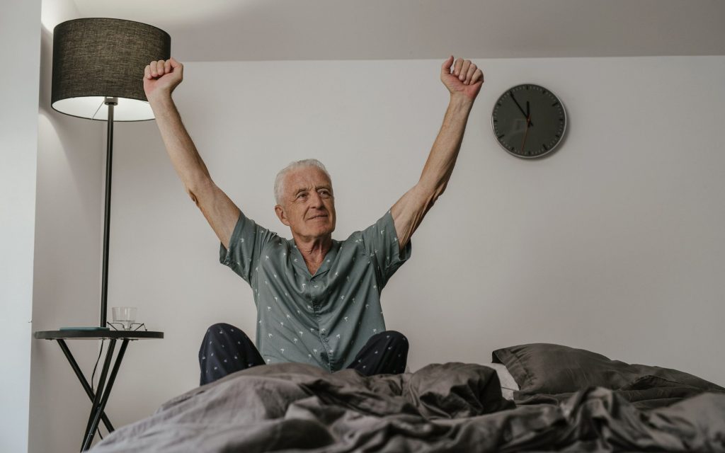 Hombre de más de 60 años estirando los brazos al despertar, sentado en la cama